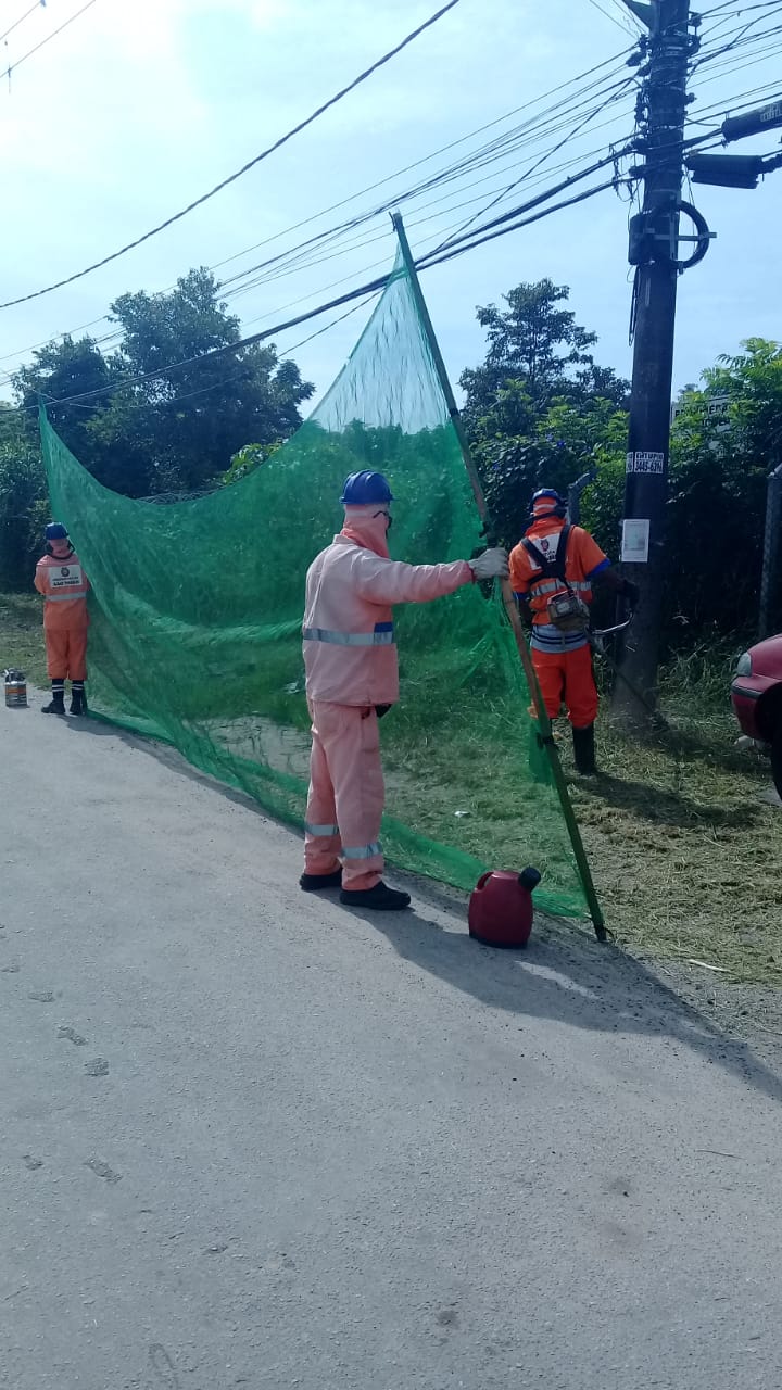 Três funcionários da Prefeitura fazem corte de mato à beira da pista, com proteção de tela verde. 